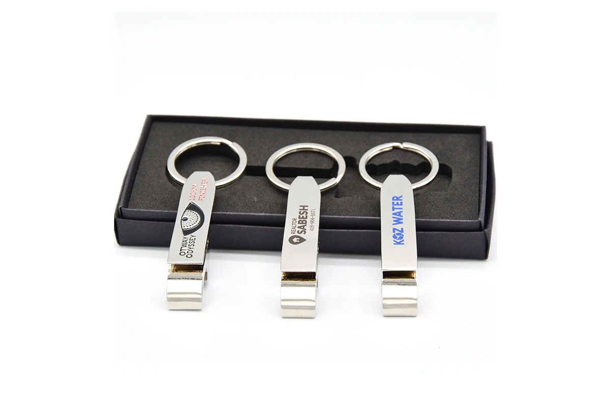 Printed logos on three silver-colored bottle opener keychains beside a foam-lined box