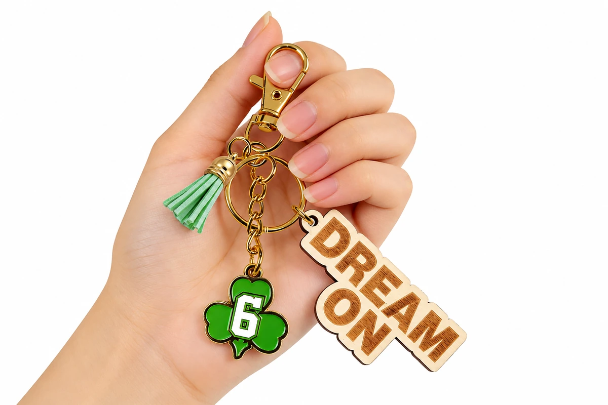 A hand holding a wooden DREAM ON keychain with a green tassel and gold-colored hardware.