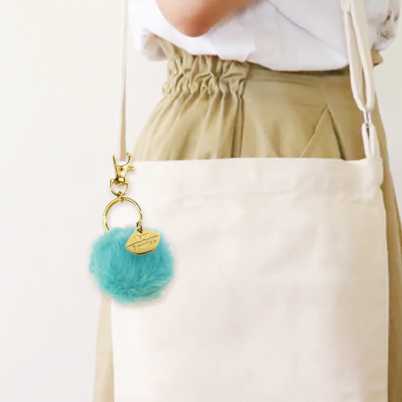 Turquoise pom-pom keychain clipped to a cream shoulder bag.
